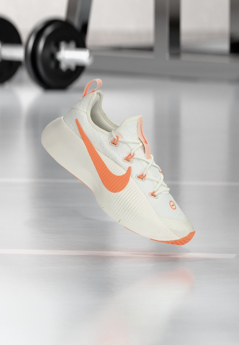 Weißer und orangefarbener Nike Laufschuh, der über einem reflektierenden Fitnessboden schwebt, mit verschwommenem Gewichtheber-Equipment im Hintergrund.