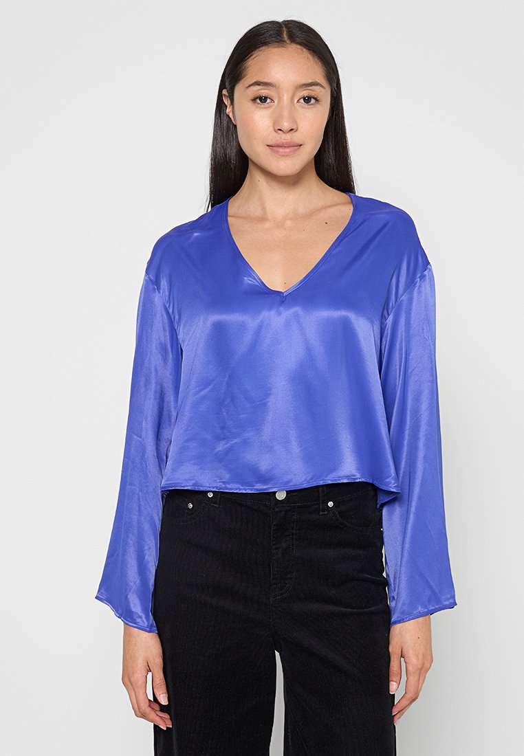V-Ausschnitt, langärmlige Bluse aus glänzendem königsblauem Stoff, cropped Design mit glatter Textur, kombiniert mit hochgeschnittenen schwarzen Cordhosen.