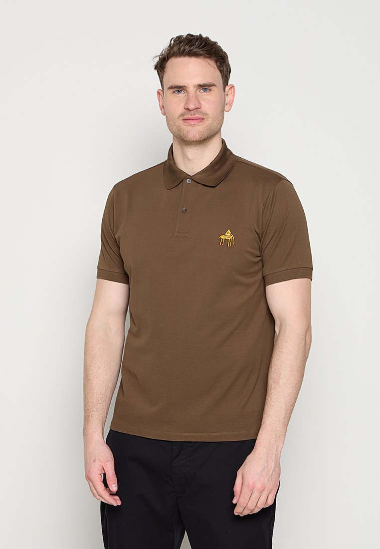 Mann trägt ein braunes kurzärmeliges Poloshirt mit einem kleinen gelben bestickten Logo auf der Brust, kombiniert mit schwarzen Hosen, steht vor einer weißen Wand.