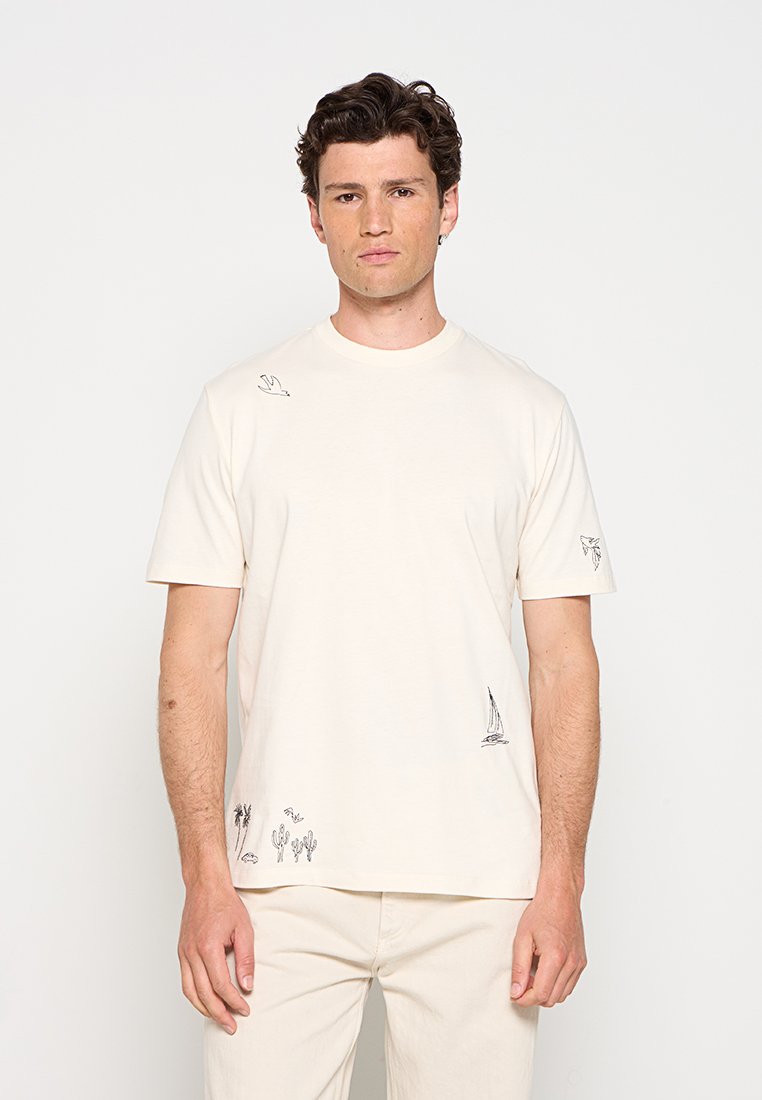 Mann trägt ein cremefarbenes T-Shirt mit kleinen schwarzen Linienzeichnungen von Vögeln, einem Segelboot, Kakteen und Pflanzen, kombiniert mit passender Hose.