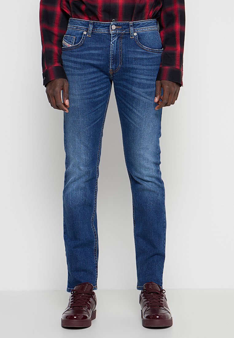 Mann trägt mittelblaue Slim-Fit-Jeans, rot-schwarz karierte Hemd und dunkelrote glänzende Turnschuhe, steht vor einem einfarbigen Hintergrund.