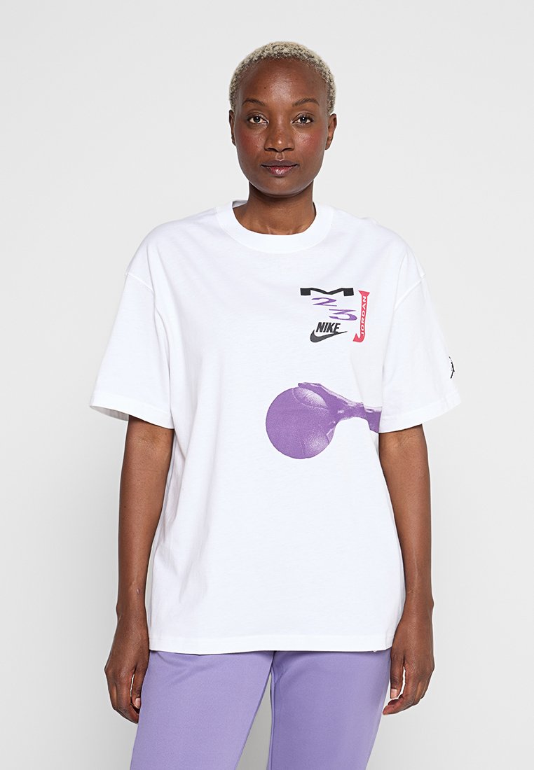 Frau mit kurzen blonden Haaren, die ein weißes Nike Jordan T-Shirt mit lila Basketball-Design und lavendelfarbene Hose vor einfarbigem Hintergrund trägt.