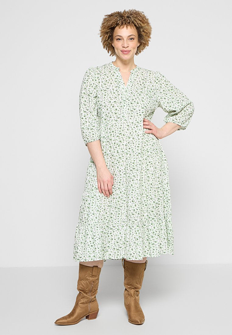 Frau mit lockigem Haar, die ein weißes Midikleid mit kleinem Blumenmuster und beigefarbene kniehohe Cowboy-Stiefel trägt, steht mit der Hand in der Hüfte.