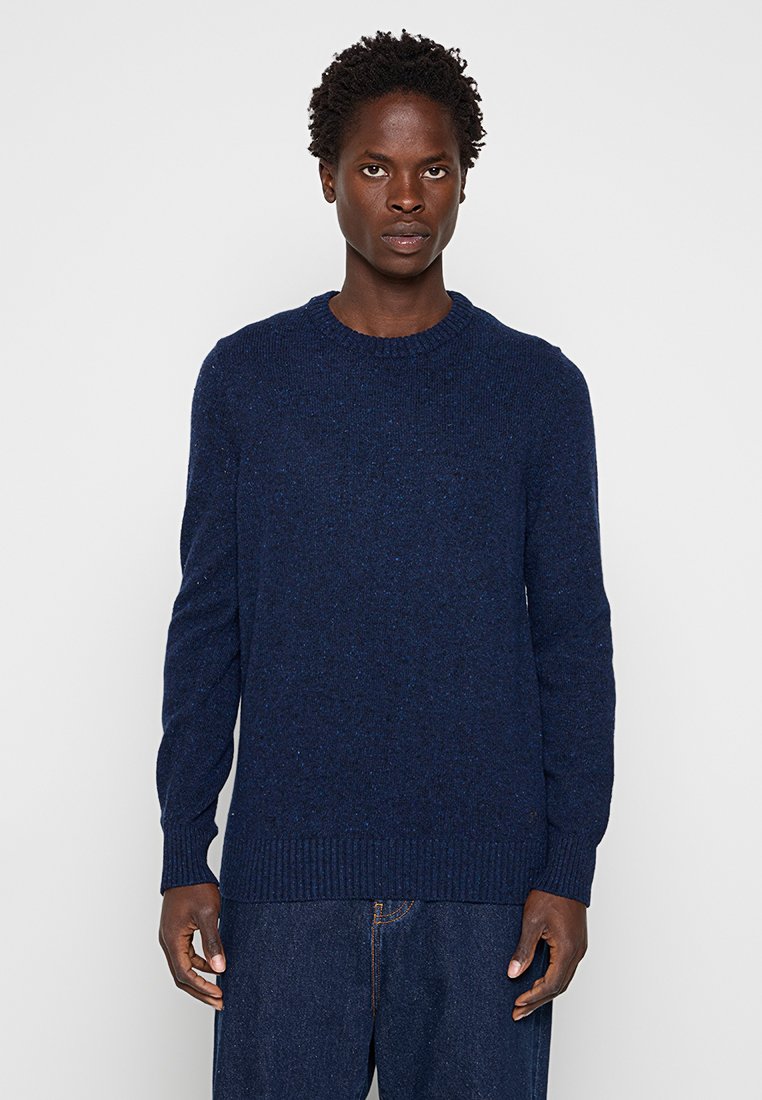 Person mit kurzen lockigen Haaren, die einen marineblauen Strickpullover und dunkelblaue Hosen trägt, steht vor einem schlichten hellen Hintergrund.