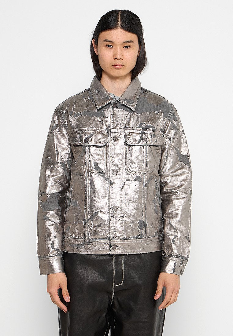 Person mit langen dunklen Haaren, die eine silberne metallische Jacke mit geknöpften Taschen und schwarze Hosen trägt, vor einem einfarbigen Hintergrund.