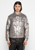 Person mit langen dunklen Haaren, die eine silberne metallische Jacke mit geknöpften Taschen und schwarze Hosen trägt, vor einem einfarbigen Hintergrund.