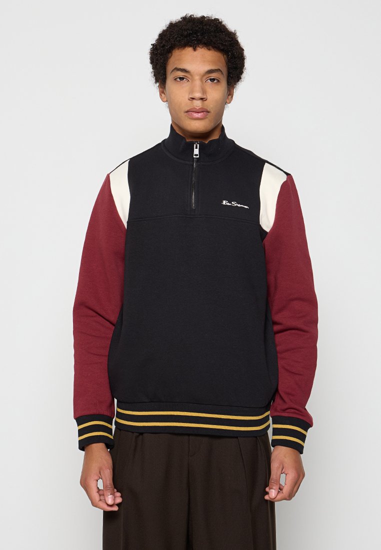 Schwarzer Sweatshirt mit Reißverschluss am Hals und kastanienbraunen Ärmeln. Hat ein cremefarbenes Paneel sowie gestreifte Rippbündchen und -saum. Logo auf der Brust in Weiß.