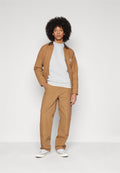 Junger Mann mit Afro-Frisur, der eine braune Jacke und Hose, ein weißes Sweatshirt und weiße Turnschuhe trägt und vor einem schlichten hellen Hintergrund steht.