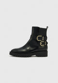 Schwarzer Leder-Stiefelette mit zwei goldenen Schnallen an den Riemen, grober Gummisohle und Zuglasche hinten vor einem schlichten hellen Hintergrund.