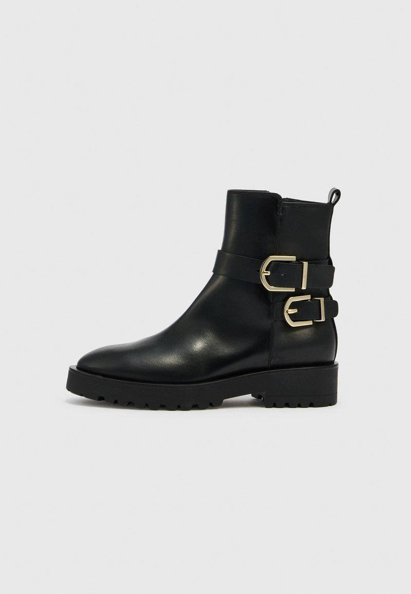 Schwarzer Leder-Stiefelette mit zwei goldenen Schnallen an den Riemen, grober Gummisohle und Zuglasche hinten vor einem schlichten hellen Hintergrund.