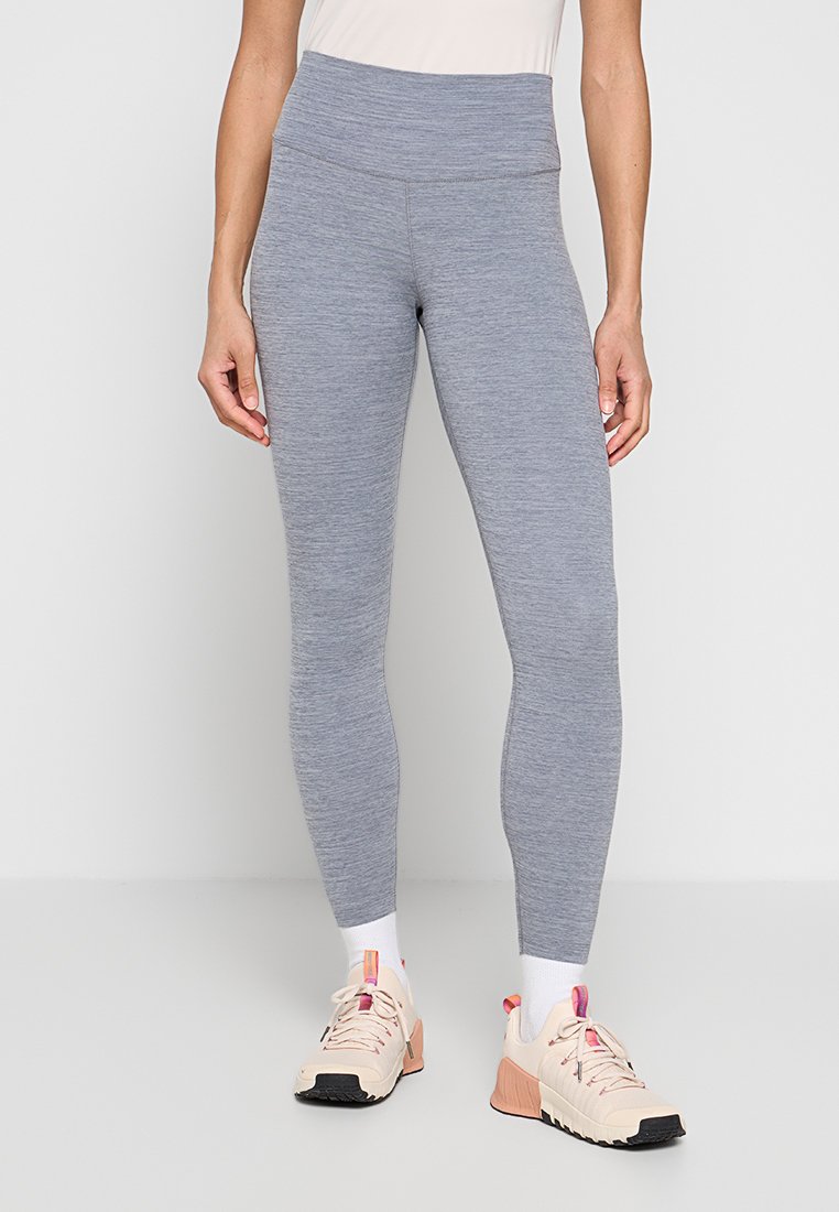 Frau trägt hochtaillierte graue Leggings mit melierter Textur, weiße Socken und beige Sportschuhe mit pinken Akzenten, steht vor neutralem Hintergrund.