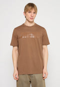 Junger Mann mit hellen Haaren, der ein braunes Diesel-T-Shirt und beige Hosen trägt, steht vor einem einfarbigen hellgrauen Hintergrund.