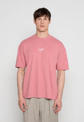 Mann, der nach vorne schaut, trägt ein lockeres pinkes T-Shirt mit kleinem weißem Text und hellbeigen Hosen vor einem einfarbigen Hintergrund.