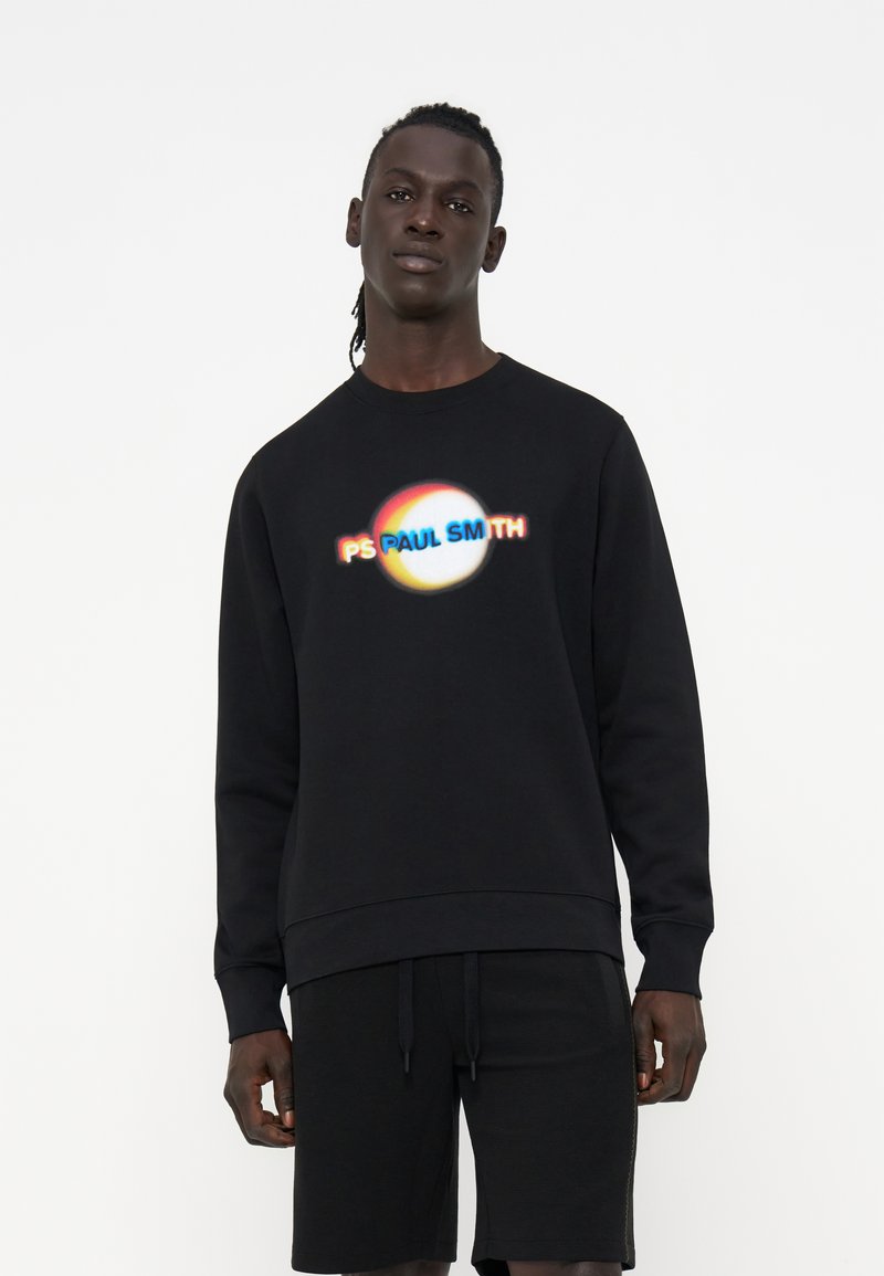 Männliches Modell trägt einen schwarzen Sweatshirt mit einem farbenfrohen verschwommenen kreisförmigen Logo und dem Text „PS PAUL SMITH“ vorne, kombiniert mit schwarzen Shorts.