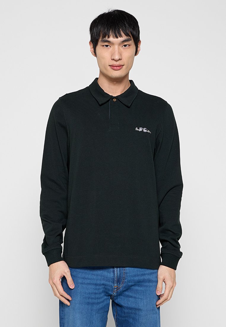 Mann trägt ein schwarzes Langarm-Poloshirt mit kleinem weißem Logo auf der Brust und blaue Jeans, steht vor schlichtem Hintergrund.