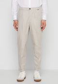 Mann trägt beige zulaufende Hose, weißes Hemd, weiße Socken und weiße Sneaker mit braunen Sohlen, steht vor einem einfarbigen Hintergrund.