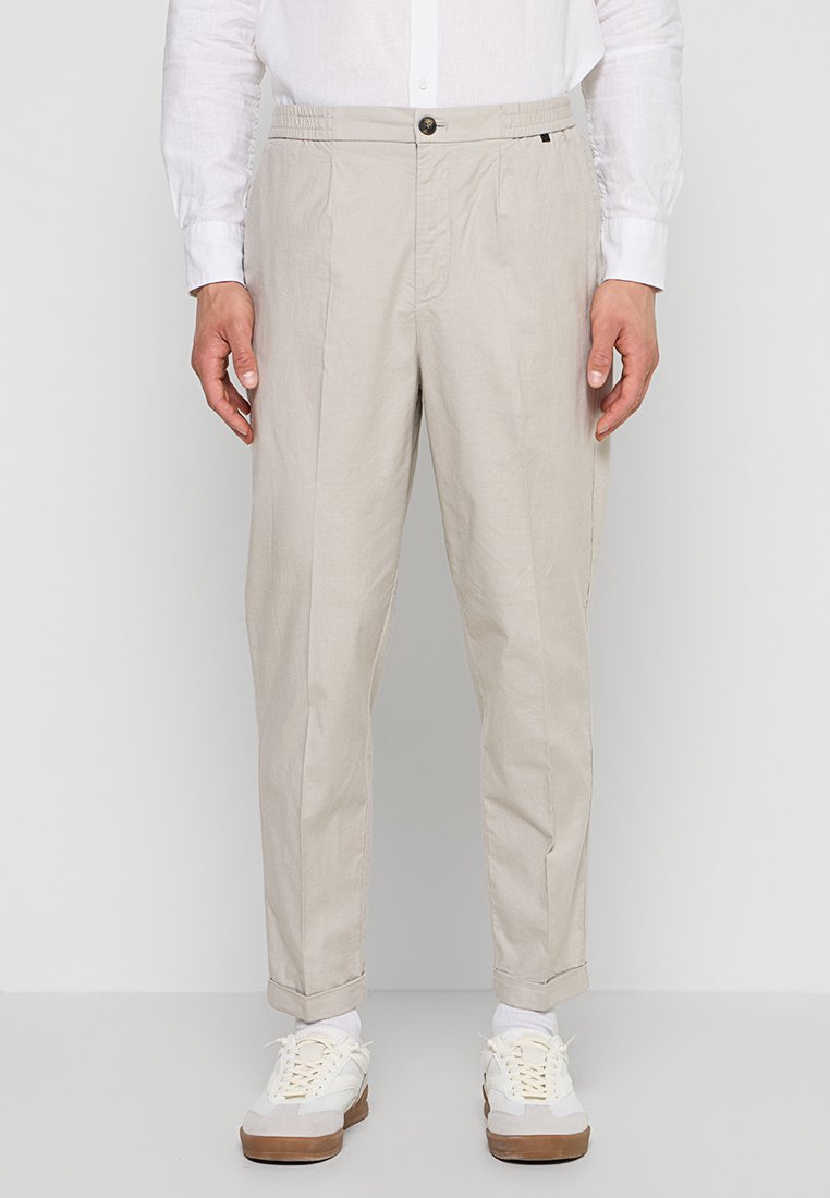 Mann trägt beige zulaufende Hose, weißes Hemd, weiße Socken und weiße Sneaker mit braunen Sohlen, steht vor einem einfarbigen Hintergrund.