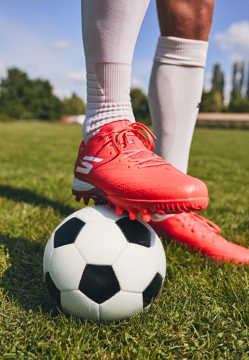 Helle rote Fußballschuhe mit strukturierten Oberflächen und weißen Akzenten, die auf einem schwarz-weißen Fußball auf grünem Gras liegen.