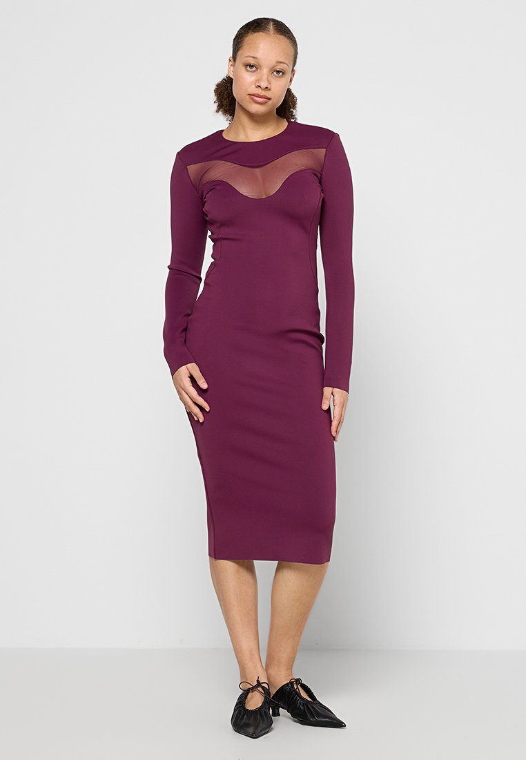 Ein figurbetontes, langärmliges Kleid in Dunkellila mit einem transparenten Einsatz am Ausschnitt, kniehoch geschnitten, kombiniert mit schwarzen Schuhen.