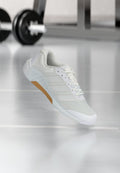 Weißer Sportschuh mit Mesh- und synthetischem Obermaterial, Gummisohlen-Detail, geschnürte Schnürsenkel, schwebend über reflektierendem Fitnessboden mit Hantel im Hintergrund.