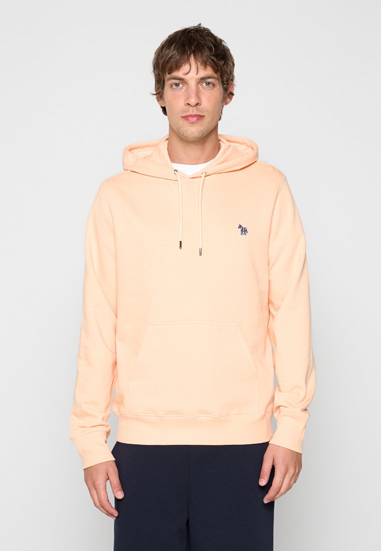 Junger Mann trägt einen pastellpfirsichfarbenen Hoodie mit kleinem Zebra-Logo auf der Brust und dunkler Hose, steht vor einem schlichten Hintergrund.