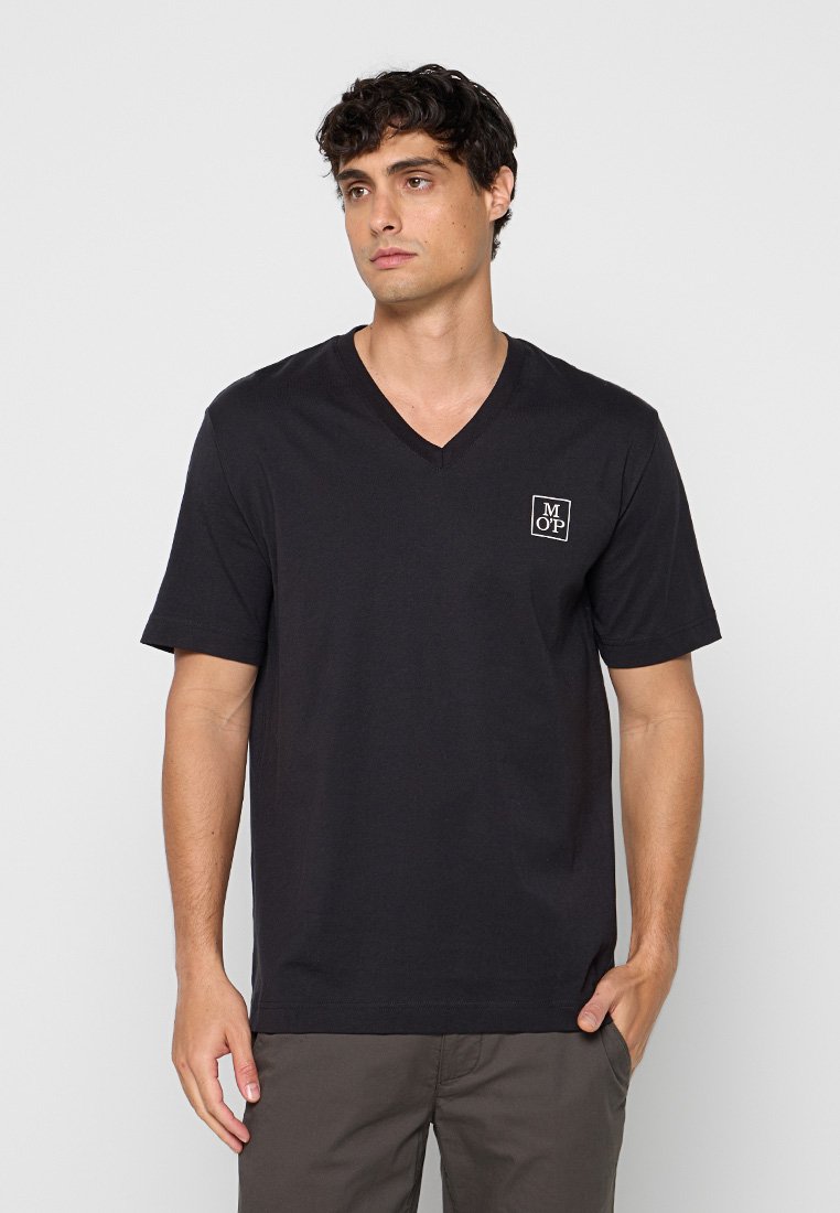Junger Mann in schwarzem V-Ausschnitt T-Shirt mit kleinem weißem Quadrat-Logo auf der linken Brust, vor einem einfarbigen hellen Hintergrund stehend.