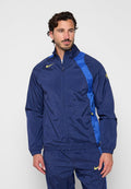 Mann trägt eine marineblaue und königsblaue Nike-Trainingsjacke mit gelben Swoosh-Logos und steht vor einem schlichten hellgrauen Hintergrund.