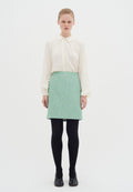 Cremfarbene Bluse mit Rundkragen und Knopfdetails, kombiniert mit einem strukturierten mintgrünen Rock, schwarzen Strumpfhosen und schwarzen Schuhen. Weißer Hintergrund.