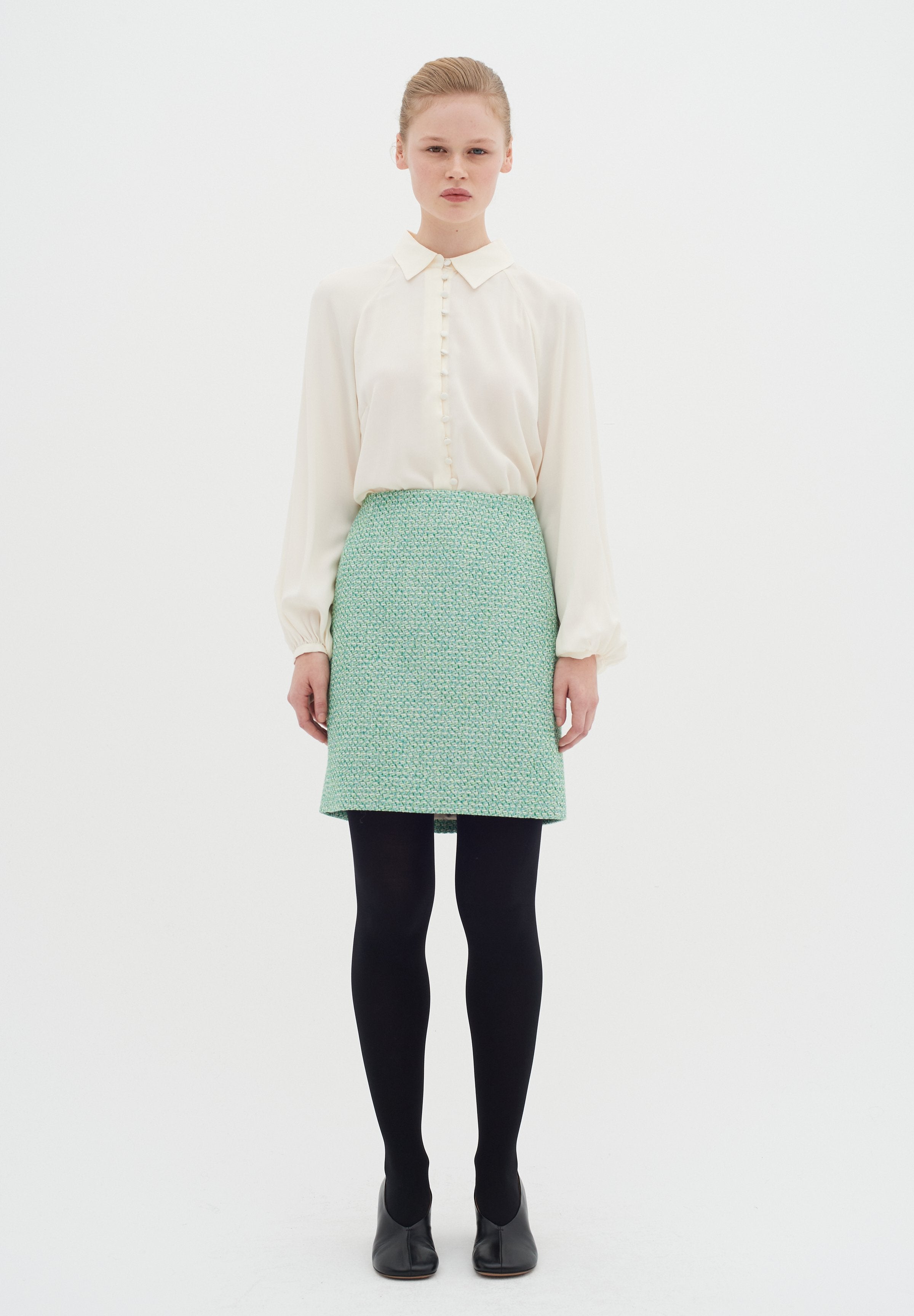 Cremfarbene Bluse mit Rundkragen und Knopfdetails, kombiniert mit einem strukturierten mintgrünen Rock, schwarzen Strumpfhosen und schwarzen Schuhen. Weißer Hintergrund.