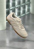 Beiger Wildledersneaker mit Gummisohle und Schnürsenkeln, mit "HUGO" geprägt, schwebend vor einer Glasbausteinwand und einem Betonboden im Hintergrund.