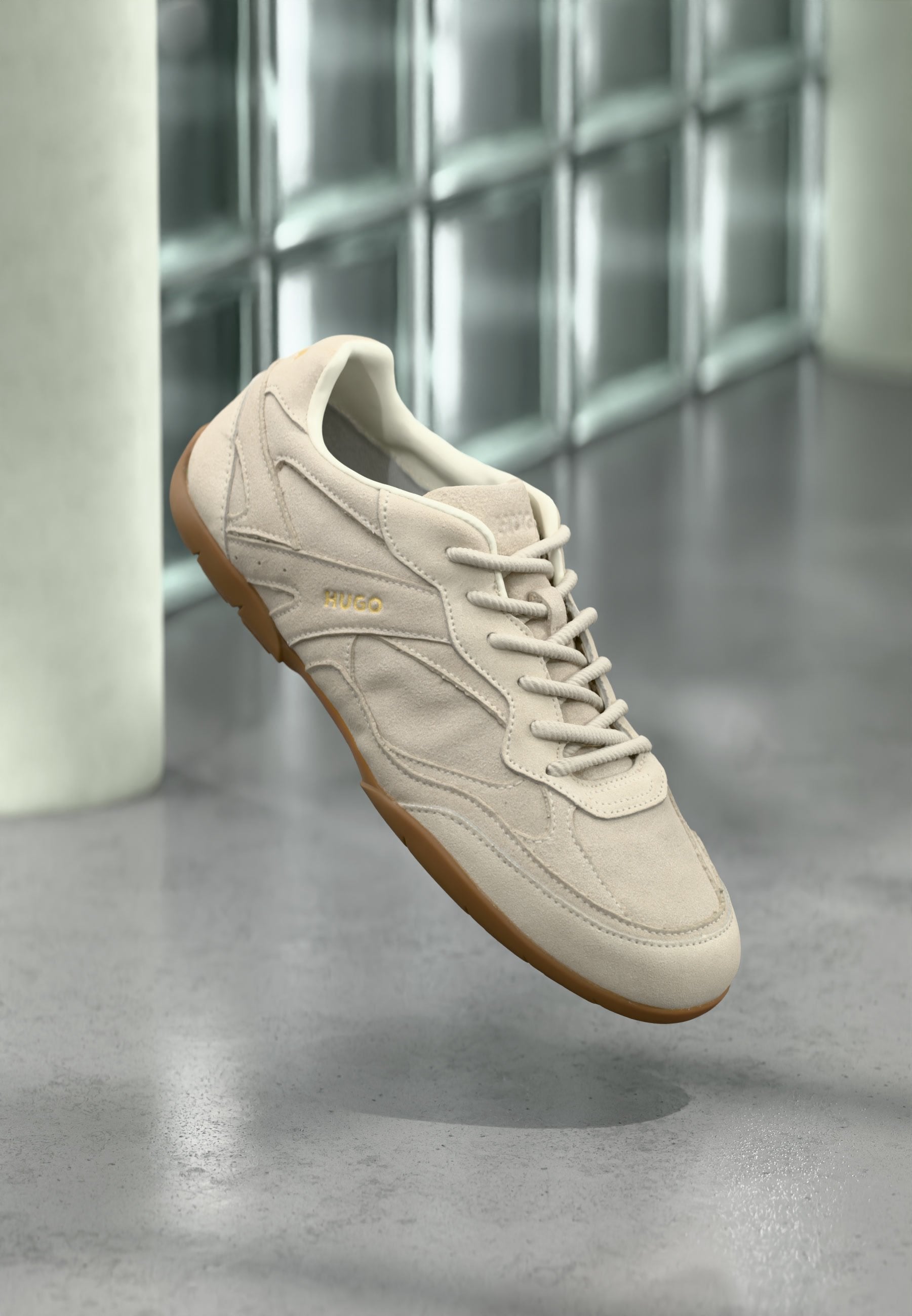 Beiger Wildledersneaker mit Gummisohle und Schnürsenkeln, mit "HUGO" geprägt, schwebend vor einer Glasbausteinwand und einem Betonboden im Hintergrund.