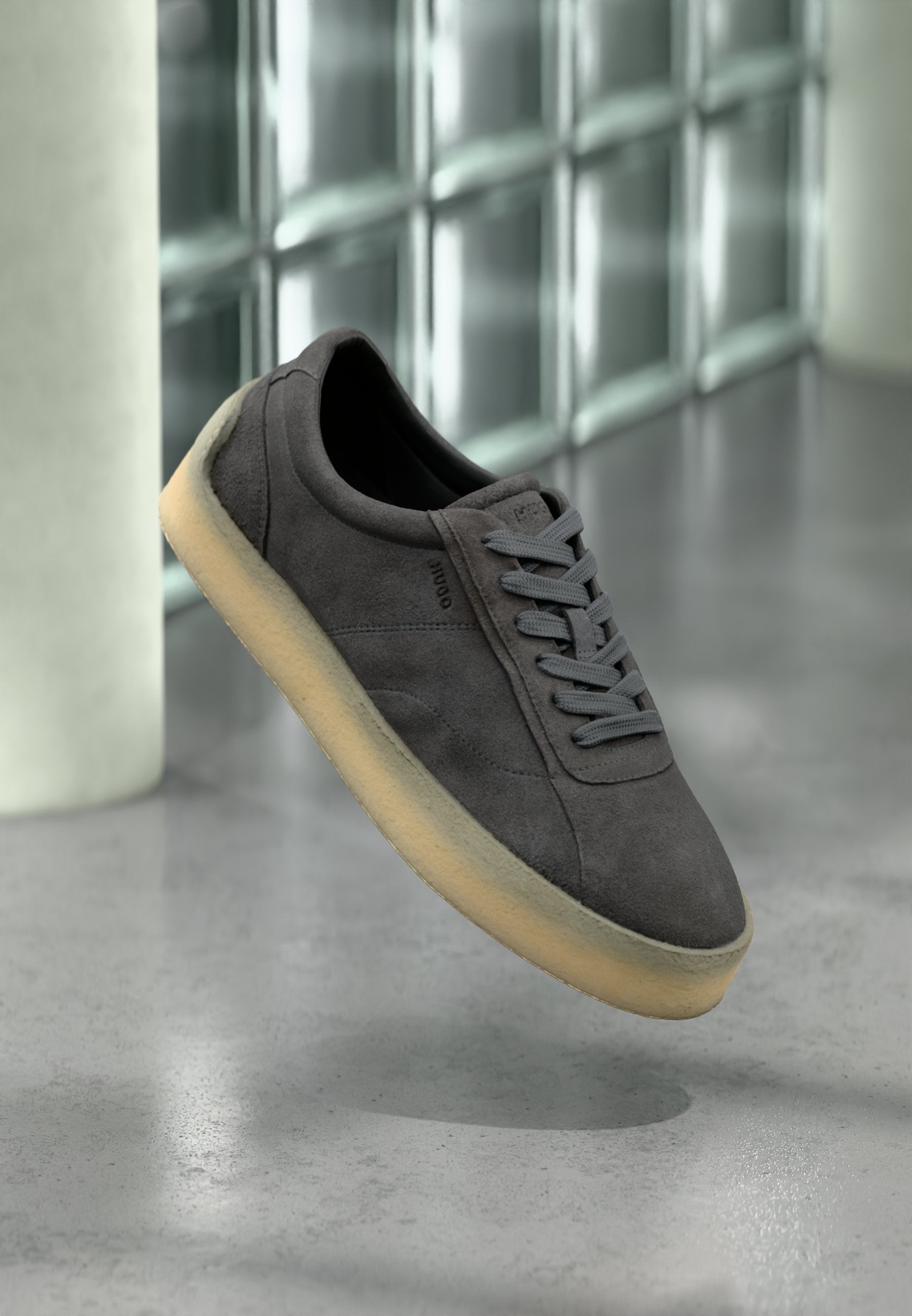 Dunkelgrauer Wildleder-Sneaker mit hellbeiger Sohle und grauen Schnürsenkeln, schwebend über einem polierten Betonboden vor einer Glasfliesenwand.