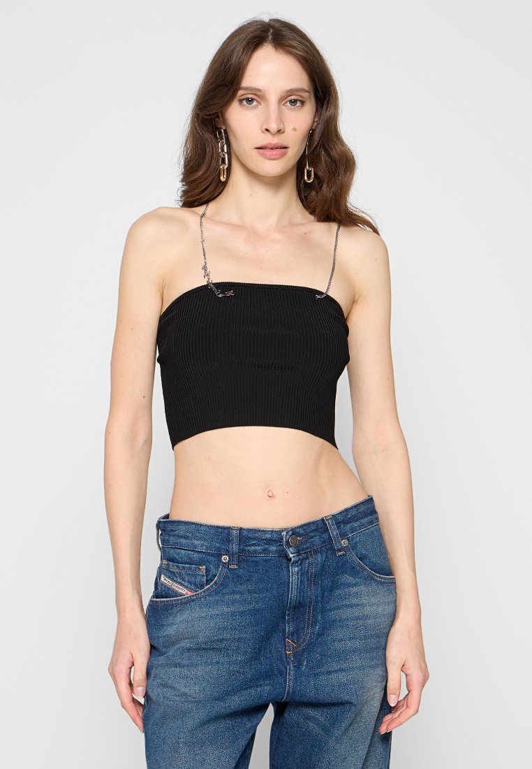 Frau mit langen braunen Haaren, die ein schwarzes trägerloses Crop-Top mit Kettenriemen und blaue lässige Jeans trägt, vor einem schlichten Hintergrund.