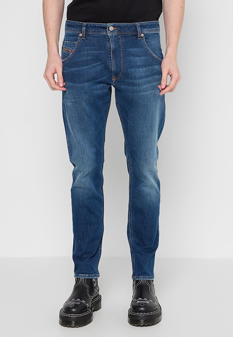Blaue Jeans im Slim-Fit mit vorderen Taschen und orangen Nähten. Kombiniert mit schwarzen Stiefeln, vor einem neutralen Hintergrund stehend.