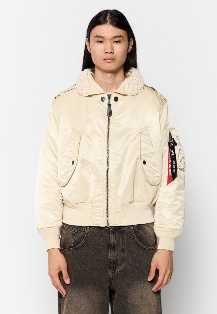 Beige Bomberjacke mit strukturiertem Kragen, Reißverschluss, zwei Fronttaschen und gerippten Bündchen. Verfügt über ein dekoratives Etikett am Ärmel.