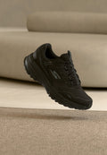 Schwarzer Sportschuh mit strukturierter Sohle und Schnürsenkeln, mit einem reflektierenden "S"-Logo, vor einem beigen Teppich und einem gebogenen Sofa im Hintergrund.