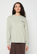 Frau mit langen, dunklen Locken, die einen hellgrünen Pullover mit „ALLSAINTS“-Logo und einen braunen Rock trägt, steht vor einem einfarbigen Hintergrund.