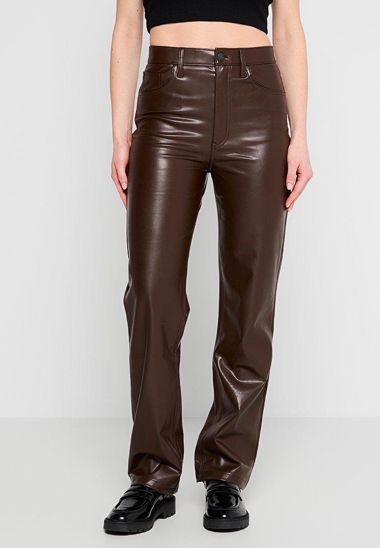 Braune Kunstlederhosen mit hoher Taille, geradem Bein, Fronttaschen und glänzender Oberfläche, kombiniert mit schwarzen, klobigen Schuhen.