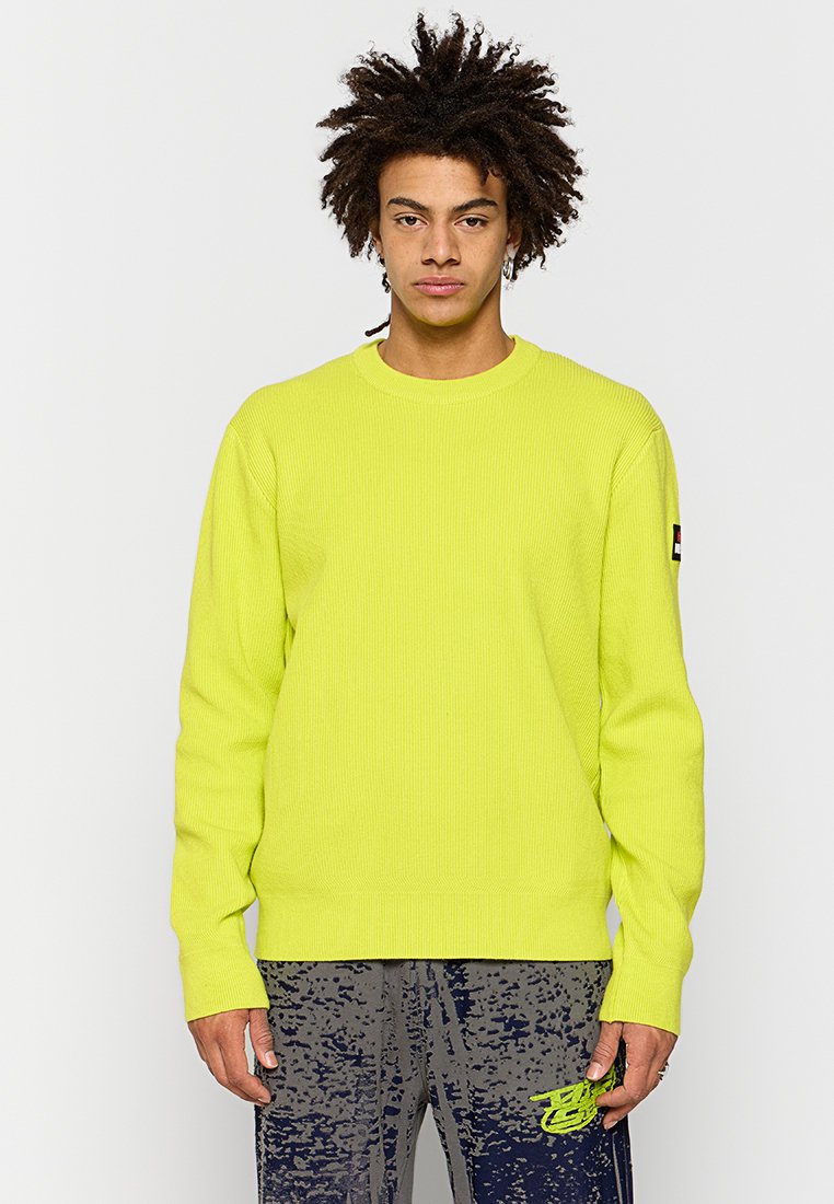 Neon-gelber, gerippter Strickpullover mit Rundhalsausschnitt und langen Ärmeln. Verfügt über ein Logo-Label am linken Ärmel. Kombiniert mit gemusterten grauen Hosen.
