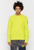 Neon-gelber, gerippter Strickpullover mit Rundhalsausschnitt und langen Ärmeln. Verfügt über ein Logo-Label am linken Ärmel. Kombiniert mit gemusterten grauen Hosen.