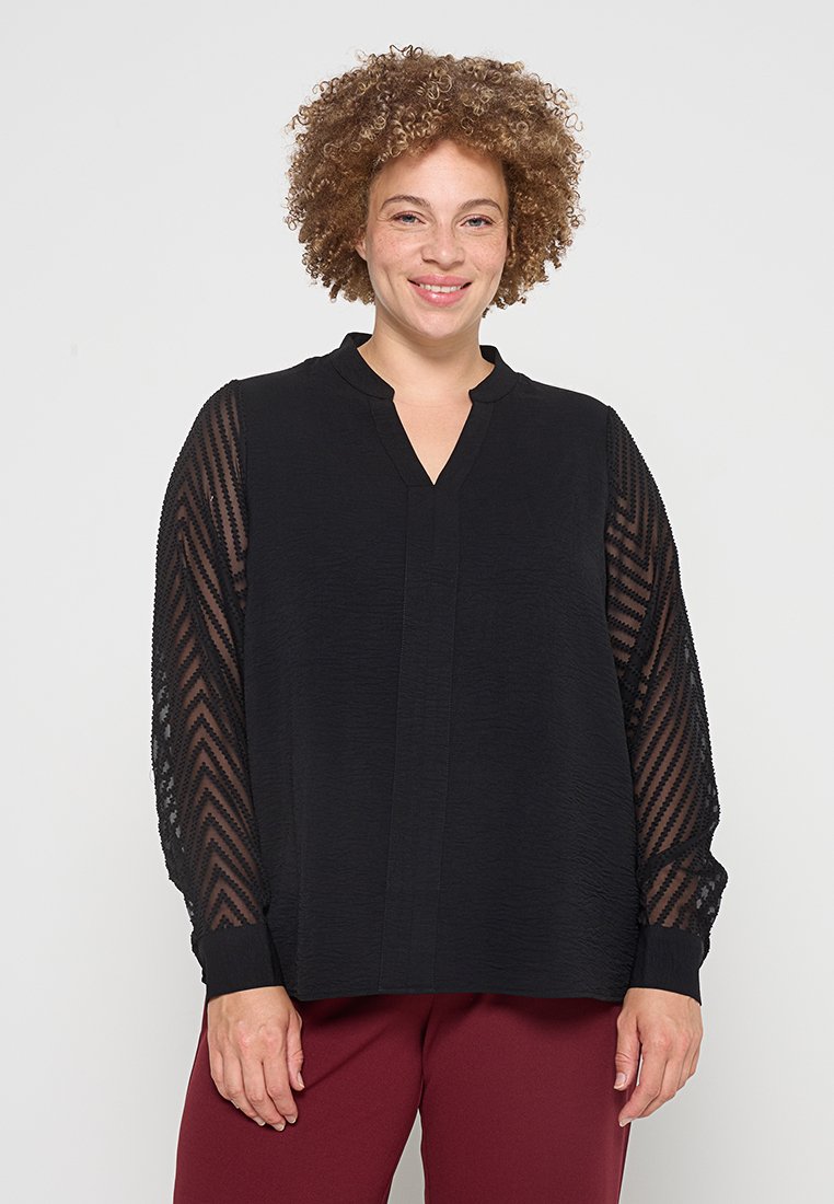 Schwarze Bluse mit strukturiertem Vorderteil und durchsichtigen, gemusterten langen Ärmeln. Mit V-Ausschnitt und Bünden an den Ärmelenden.