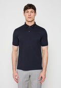Junger Mann, der ein marineblaues Polohemd mit kurzen Ärmeln und hellgraue Hose trägt, vor einem schlichten weißen Hintergrund stehend.