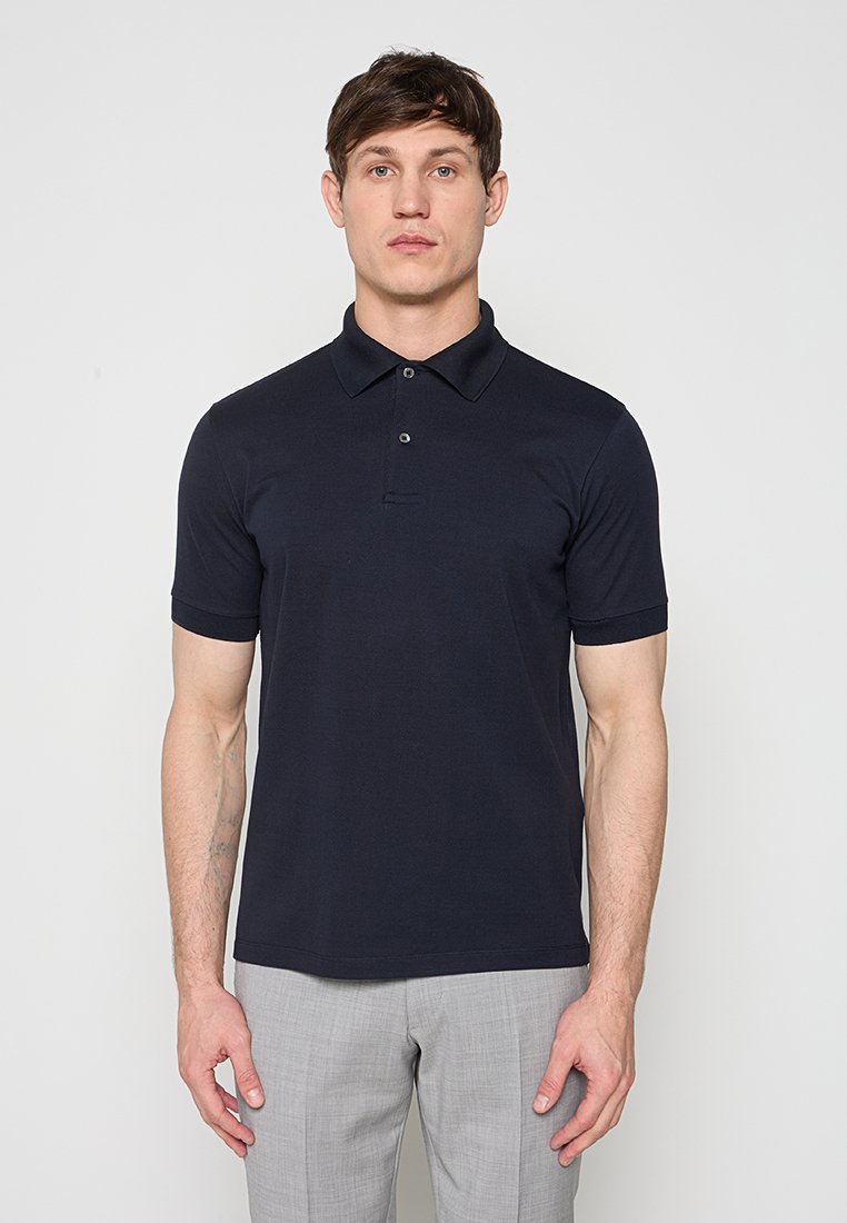 Junger Mann, der ein marineblaues Polohemd mit kurzen Ärmeln und hellgraue Hose trägt, vor einem schlichten weißen Hintergrund stehend.