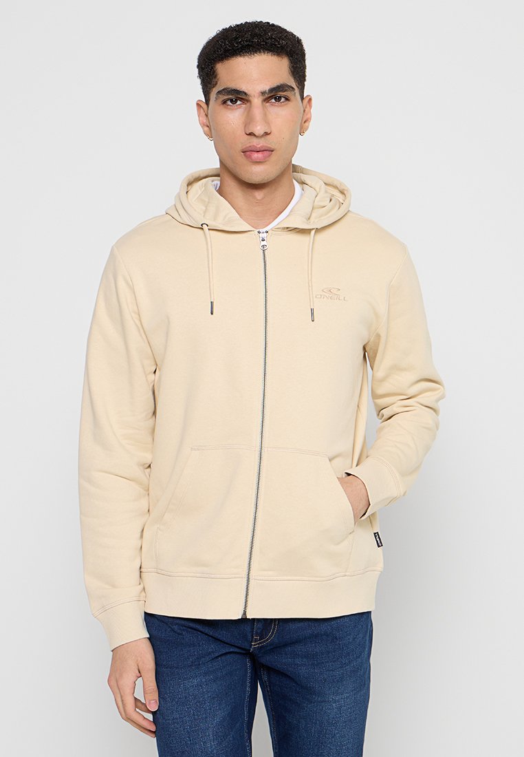 Beige Kapuzenpulli mit Reißverschluss aus weichem Stoff mit einem weißen Innenfutter, Kapuze mit Kordelzug, Fronttaschen und dezentem Logo auf der Brust.