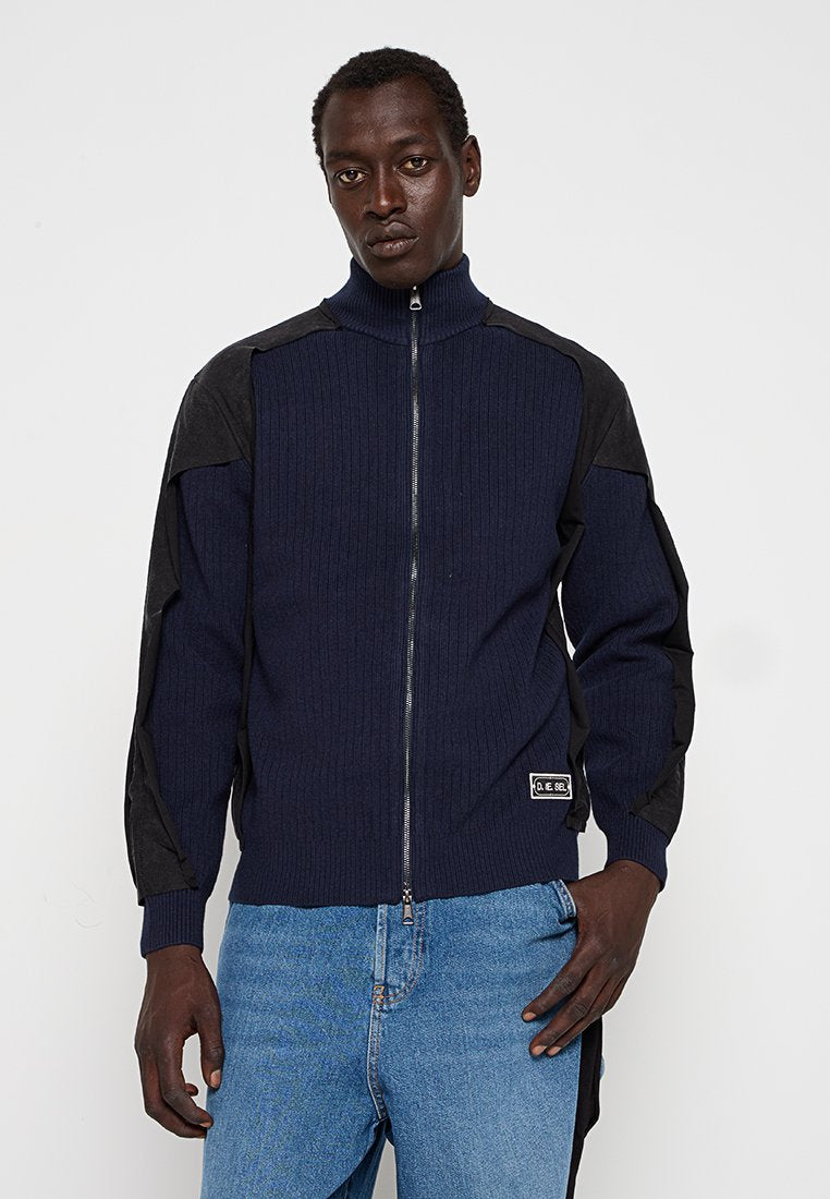 Marineblaue gerippte Zip-Up-Strickjacke mit schwarzen Panelelementen, hohem Kragen und Logo-Patch, kombiniert mit hellblauen Jeans.