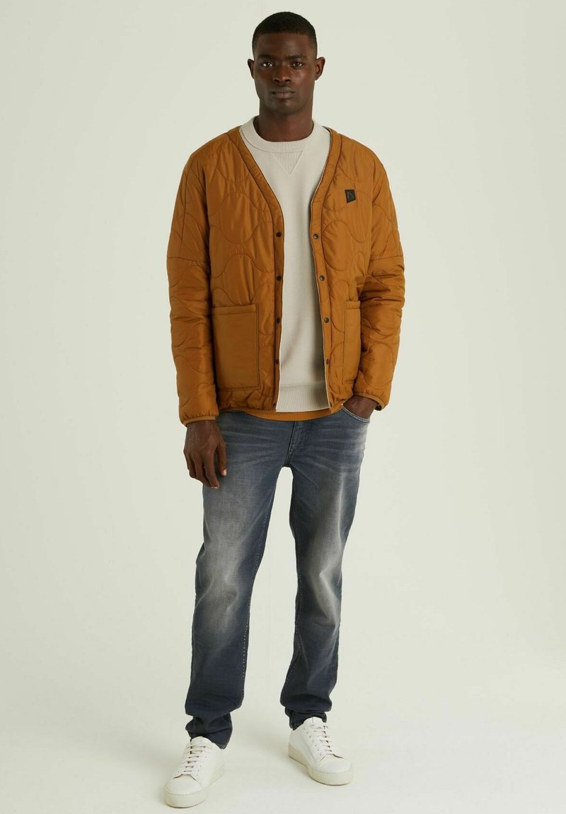 Junger Mann in brauner Steppjacke über beigem Pullover, mit verwaschenen blauen Jeans und weißen Sneakern, stehend vor einem schlichten Hintergrund.