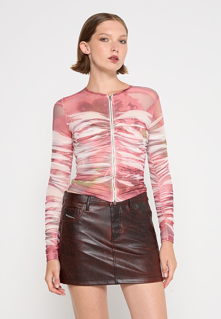 Frau in einem pink-weißen, gerafften Langarm-Zip-Up-Oberteil und einem dunkelbraunen Leder-Minirock, die vor einem schlichten Hintergrund steht.