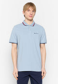 Junger Mann mit hellen Haaren, der ein hellblaues Poloshirt mit roten und marineblauen Akzenten trägt, steht vor einem schlichten weißen Hintergrund.