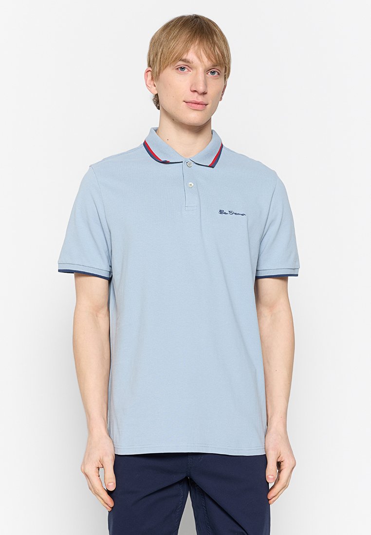 Junger Mann mit hellen Haaren, der ein hellblaues Poloshirt mit roten und marineblauen Akzenten trägt, steht vor einem schlichten weißen Hintergrund.