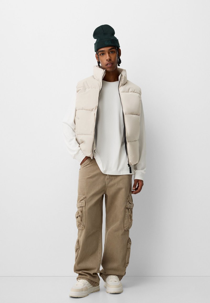 Beige Puffer-Weste über einem weißen Langarmshirt, kombiniert mit beigefarbenen Cargo-Hosen und cremefarbenen Sneakers. Eine dunkle Mütze und strukturierte Haare setzen Akzente.